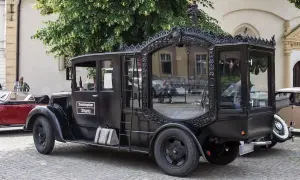 Czarny, zabytkowy karawan z ozdobnymi ramami okiennymi i napisem "Bestattungshaus Rogenz". Obok stoi czerwony kabriolet.