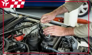 Mechanik naprawia silnik samochodu, używając klucza nasadowego. W tle widać logo firmy, przypominające kształtem nagar.