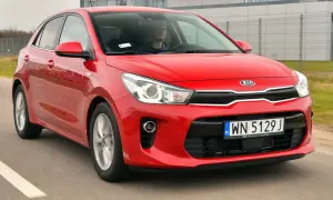 Czerwony kia rio 4 w ruchu, z kierowcą za kierownicą, na tle nowoczesnego budynku.