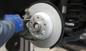 Mechanik w niebieskiej rękawicy dokręca śrubę podczas wymiany tarcz hamulcowych. Widać nowy, lśniący dysk hamulcowy i sprężynę amortyzatora.