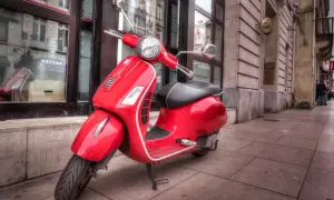 Czerwony skuter Vespa, idealny do zdobycia prawa jazdy AM, stoi na chodniku przed budynkiem.