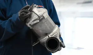 Ręce w rękawiczkach trzymają metalowy element, prawdopodobnie katalizator samochodowy, z widocznym napisem "fap".