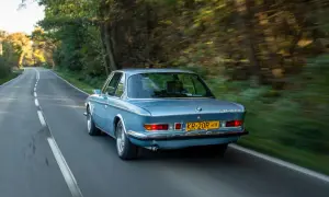 Niebieskie BMW E9 z lat 70. sunie po leśnej drodze, tworząc wrażenie prędkości.