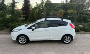 Biały Ford Fiesta z bagażnikiem dachowym, idealny do wynajem długoterminowy z wykupem. Zaparkowany na podjeździe z kostki brukowej, otoczony zielenią.