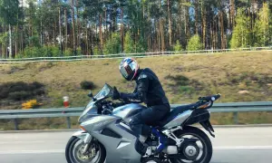Motocyklista w kasku jedzie na srebrnym Honda VFR 800 wzdłuż leśnej drogi.