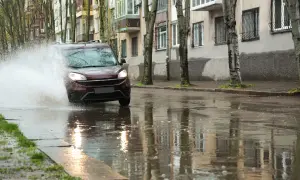 Ciemnobordowy samochód wpada w kałużę, powodując potężny rozbryzg wody. Niebezpieczny aquaplaning na mokrej ulicy.