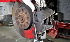 Mechanik w białych rękawiczkach dokonuje wymiany klocków hamulcowych, używając śrubokręta do podważenia starego klocka.