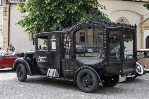 Czarny, zabytkowy karawan z ozdobnymi ramami okiennymi i napisem "Bestattungshaus Rogenz". Obok stoi czerwony kabriolet.