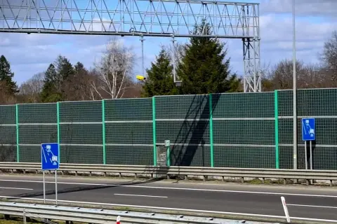Znaki informują o odcinkowym pomiarze prędkości. Za zieloną barierą dźwiękochłonną widać drzewa i niebo.