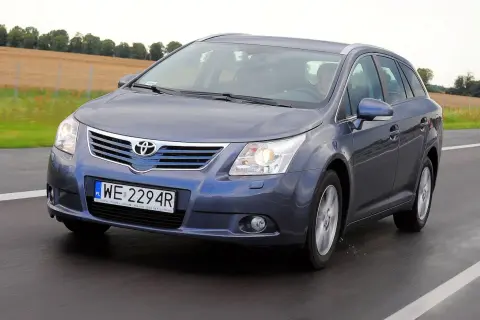 Szary kombi Toyota Avensis T27 1.8 benzyna jedzie po drodze, za nim pola i drzewa.