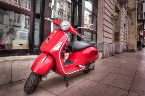 Czerwony skuter Vespa, idealny do zdobycia prawa jazdy AM, stoi na chodniku przed budynkiem.