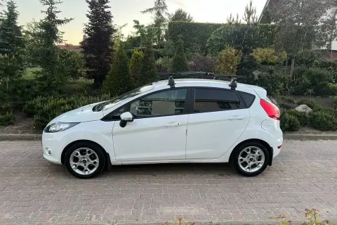 Biały Ford Fiesta z bagażnikiem dachowym, idealny do wynajem długoterminowy z wykupem. Zaparkowany na podjeździe z kostki brukowej, otoczony zielenią.