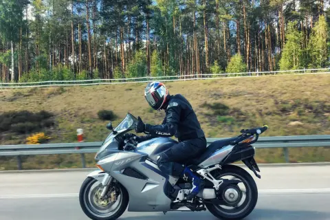 Motocyklista w kasku jedzie na srebrnym Honda VFR 800 wzdłuż leśnej drogi.