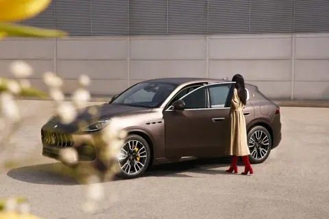 Elegancka kobieta w beżowej sukience i czerwonych kozakach wsiada do brązowego Maserati SUV.