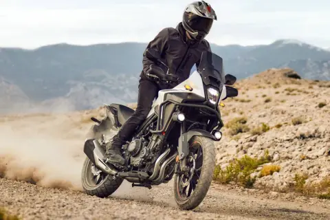 Motocyklista w kasku i kurtce pędzi na motocyklu Honda CB500X po piaszczystej drodze, wzbijając tumany kurzu.
