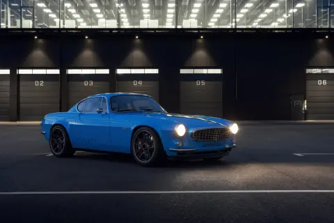 Niebieskie Volvo P1800 z nowoczesnymi felgami stoi przed budynkiem z numerowanymi bramami.