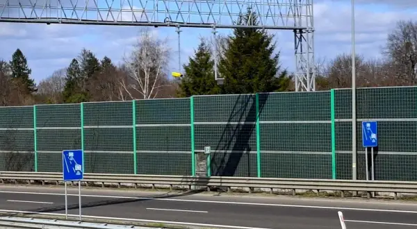 Znaki informują o odcinkowym pomiarze prędkości. Za zieloną barierą dźwiękochłonną widać drzewa i niebo.