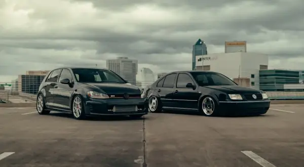 Dwa czarne Volkswageny: sportowy hatchback GTI i sedan, zaparkowane na dachu parkingu z widokiem na miasto.