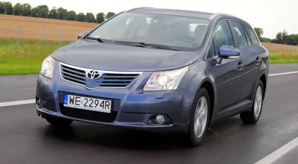 Szary kombi Toyota Avensis T27 1.8 benzyna jedzie po drodze, za nim pola i drzewa.