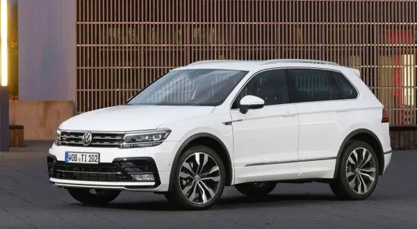 Biały Volkswagen Tiguan 2019 z eleganckimi felgami stoi przed nowoczesnym budynkiem z drewnianą fasadą.