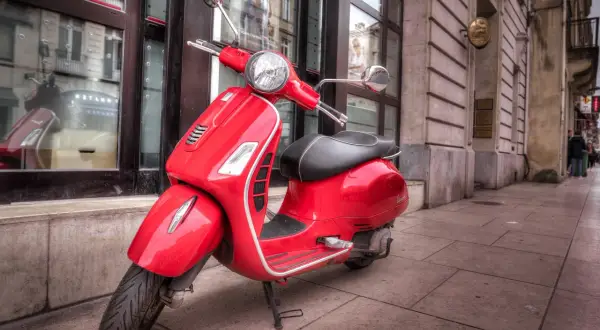 Czerwony skuter Vespa, idealny do zdobycia prawa jazdy AM, stoi na chodniku przed budynkiem.