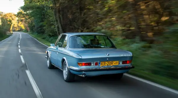 Niebieskie BMW E9 z lat 70. sunie po leśnej drodze, tworząc wrażenie prędkości.