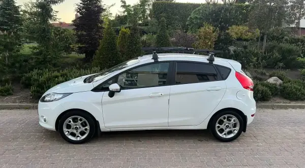 Biały Ford Fiesta z bagażnikiem dachowym, idealny do wynajem długoterminowy z wykupem. Zaparkowany na podjeździe z kostki brukowej, otoczony zielenią.