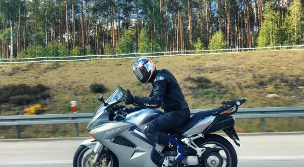 Motocyklista w kasku jedzie na srebrnym Honda VFR 800 wzdłuż leśnej drogi.