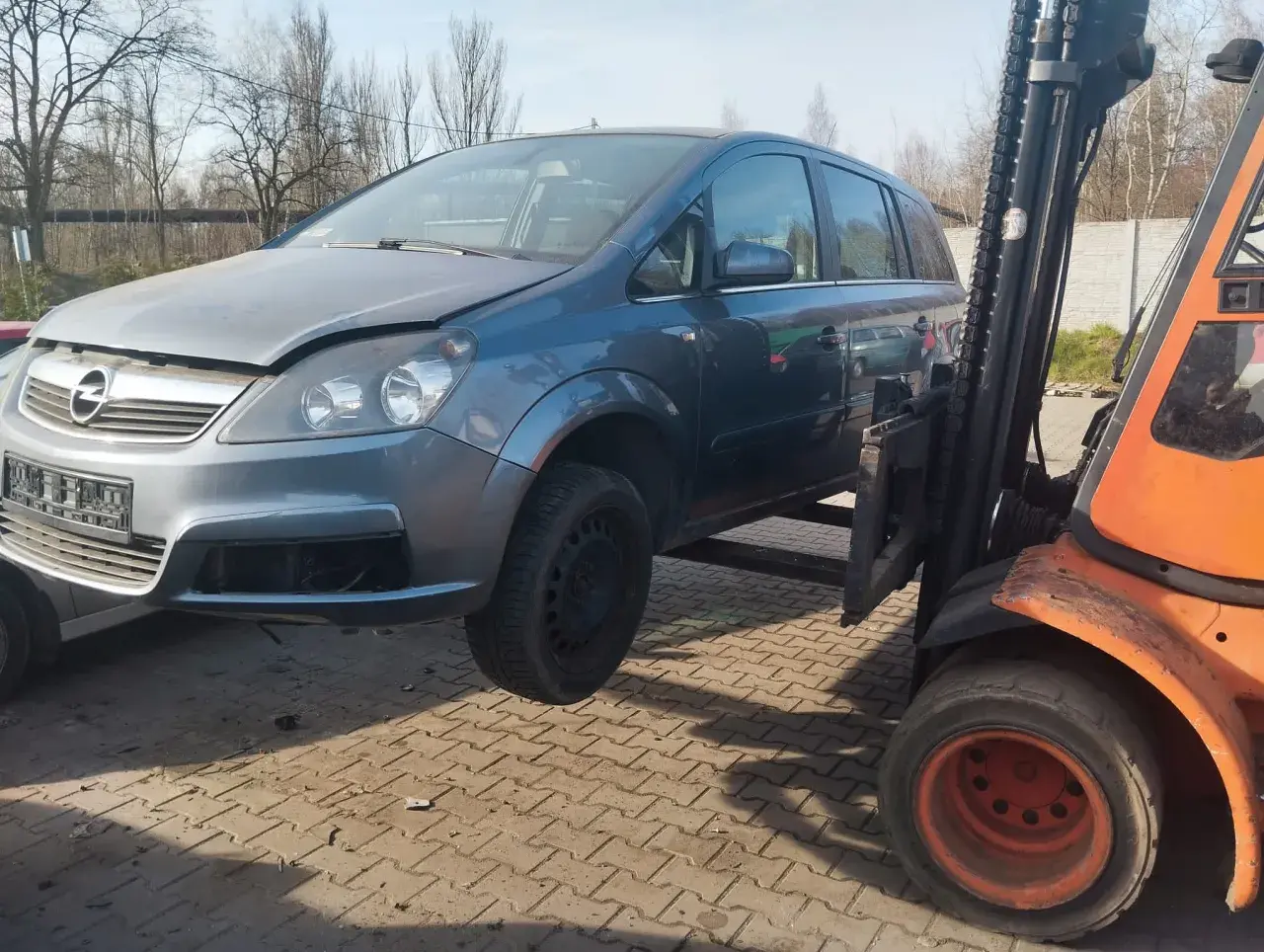 Szary Opel Zafira na złomowisku, podnoszony przez wózek widłowy. Dowiedz się, ile kosztuje złomowanie auta.