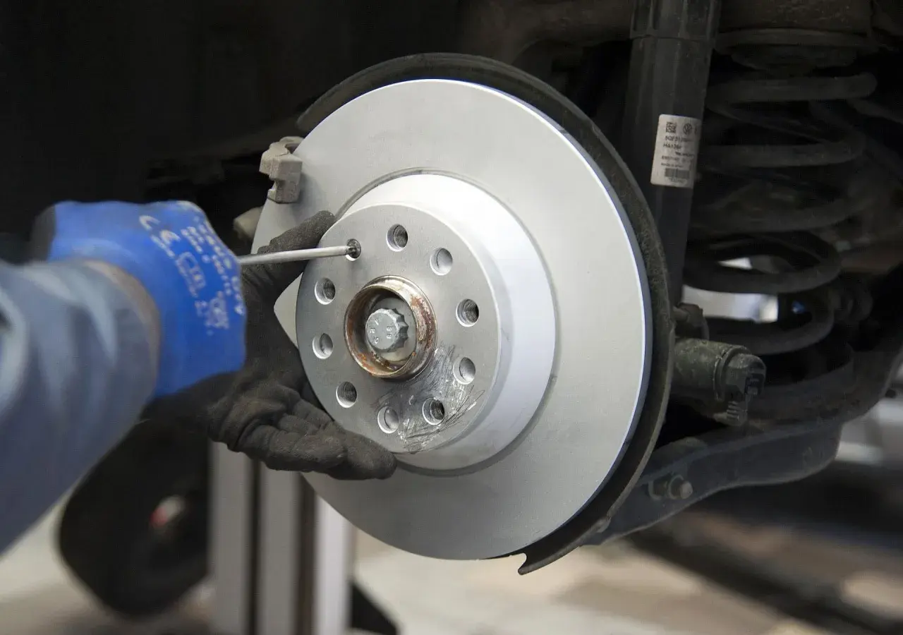 Mechanik w niebieskiej rękawicy dokręca śrubę podczas wymiany tarcz hamulcowych. Widać nowy, lśniący dysk hamulcowy i sprężynę amortyzatora.