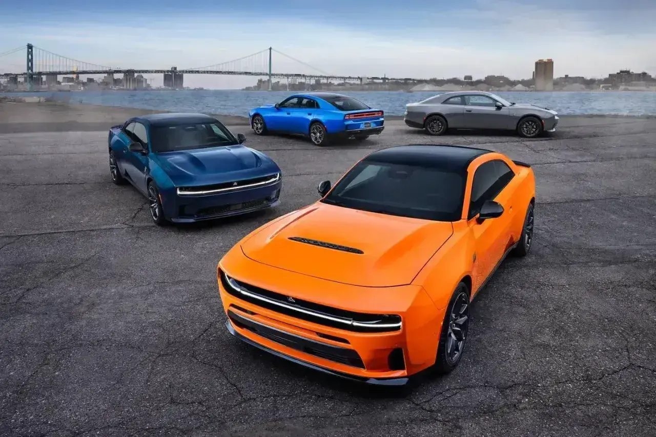Pomarańczowy i niebieski Dodge Charger Hellcat z 2024 roku, obok szarego coupe, na tle mostu i miasta.