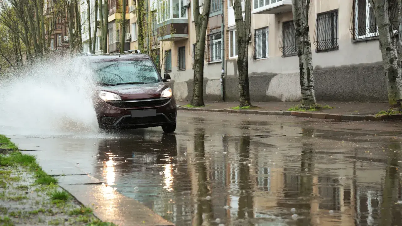 Ciemnobordowy samochód wpada w kałużę, powodując potężny rozbryzg wody. Niebezpieczny aquaplaning na mokrej ulicy.
