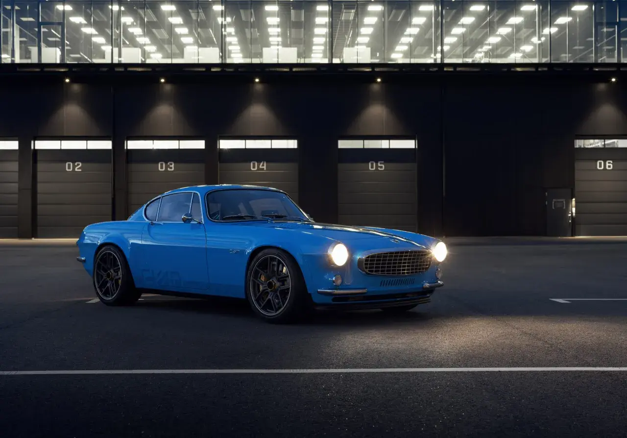 Niebieskie Volvo P1800 z nowoczesnymi felgami stoi przed budynkiem z numerowanymi bramami.