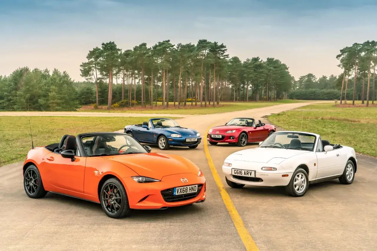 Cztery Mazdy MX-5, w tym pomarańczowy roadster, prezentują ewolucję kultowego roadstera na tle sosnowego lasu.