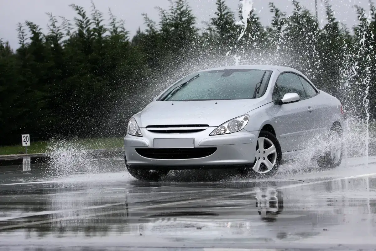 Srebrny samochód wpada w aquaplaning, rozpryskując wodę na mokrej drodze.