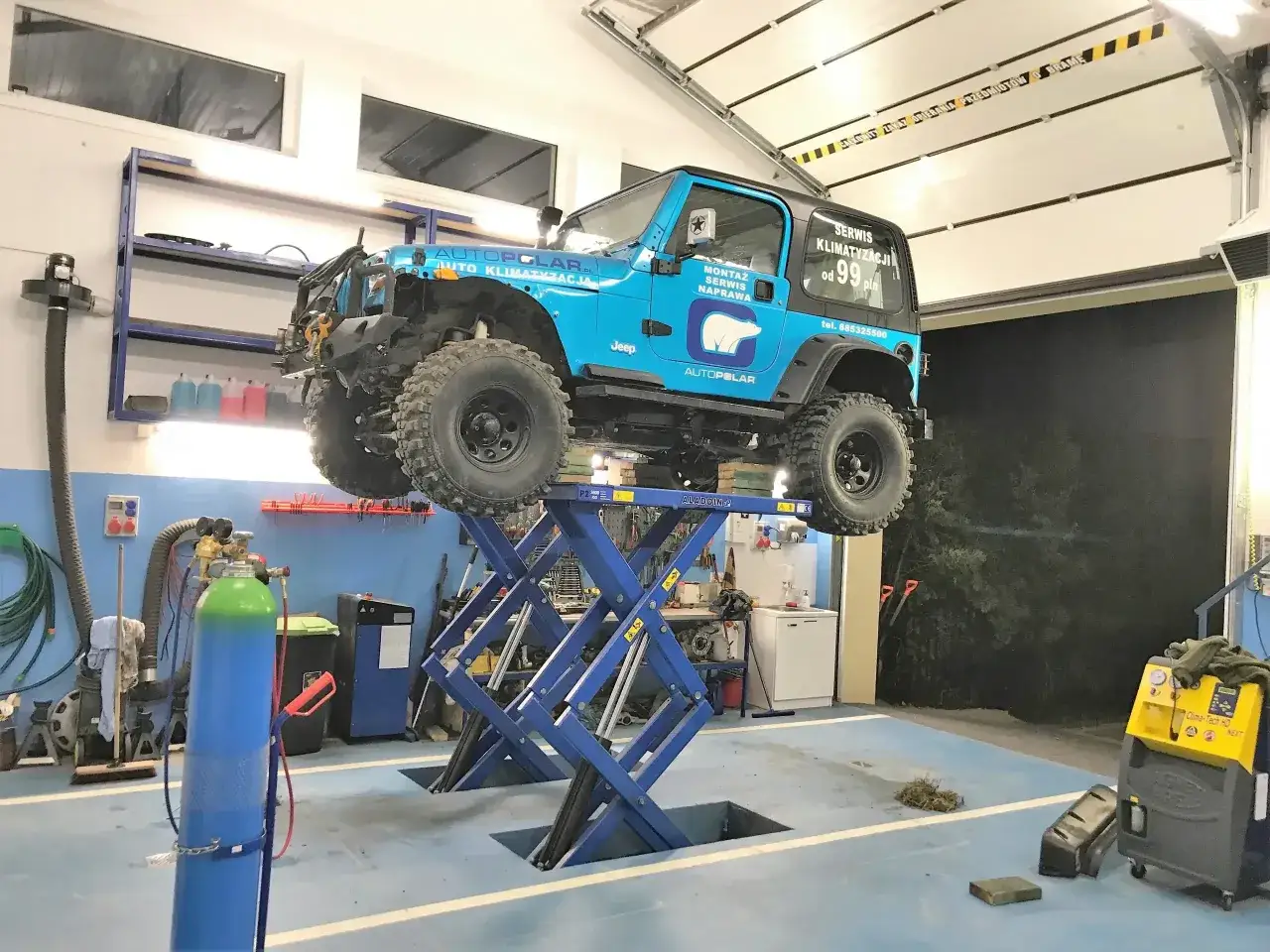Niebieski Jeep na podnośniku w warsztacie. Na drzwiach napis
