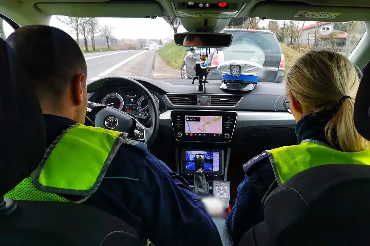 Policjanci w radiowozie, jadący drogą. Czy trzeba mieć prawo jazdy przy sobie? Na desce rozdzielczej czapka policyjna.