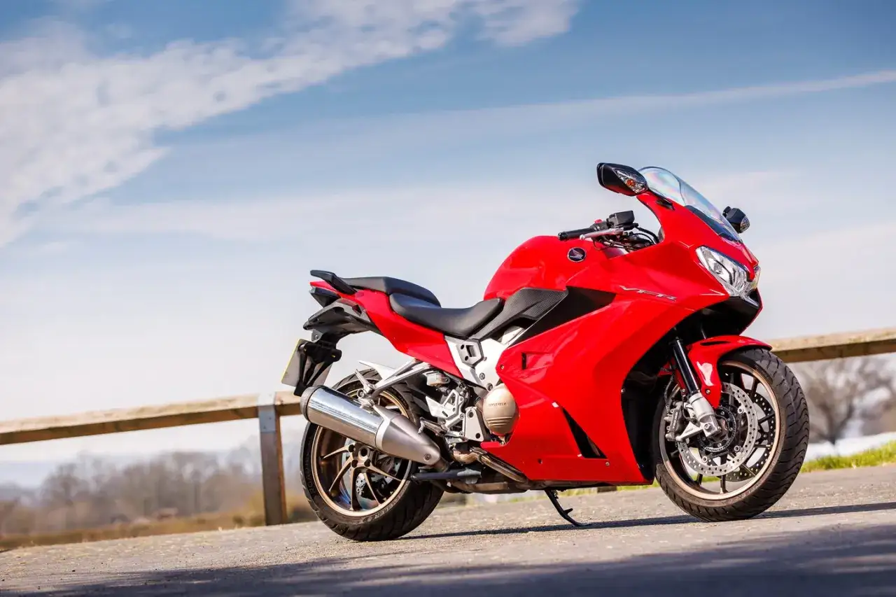 Czerwona Honda VFR 800 na tle błękitnego nieba z chmurami. Motocykl stoi na drodze, obok drewnianego płotka.