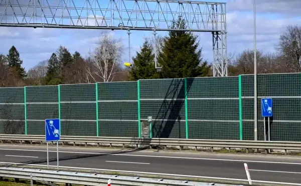 Znaki informują o odcinkowym pomiarze prędkości. Za zieloną barierą dźwiękochłonną widać drzewa i niebo.