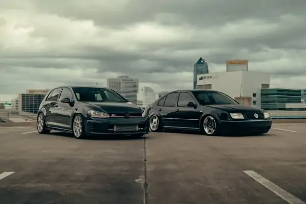 Dwa czarne Volkswageny: sportowy hatchback GTI i sedan, zaparkowane na dachu parkingu z widokiem na miasto.