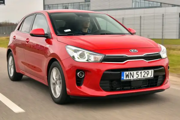 Czerwony kia rio 4 w ruchu, z kierowcą za kierownicą, na tle nowoczesnego budynku.