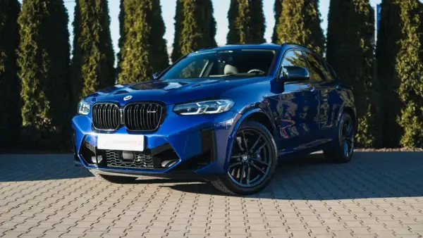 Błękitny suv BMW X4 M Competition błyszczy na tle zielonych drzew.