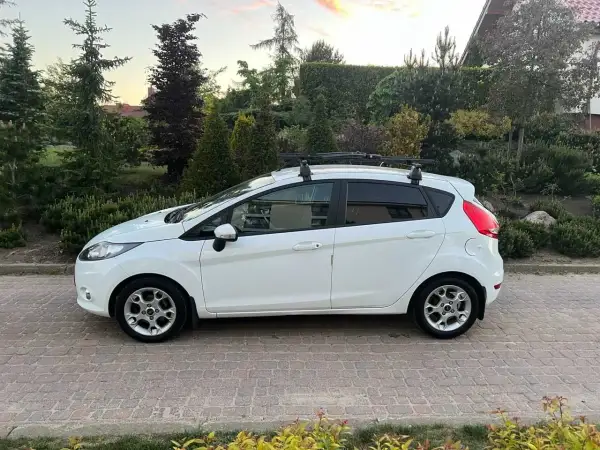 Biały Ford Fiesta z bagażnikiem dachowym, idealny do wynajem długoterminowy z wykupem. Zaparkowany na podjeździe z kostki brukowej, otoczony zielenią.