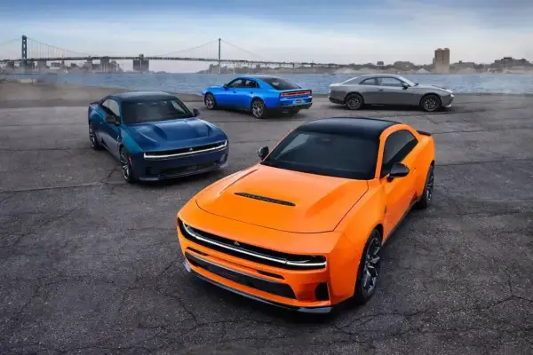 Pomarańczowy i niebieski Dodge Charger Hellcat z 2024 roku, obok szarego coupe, na tle mostu i miasta.
