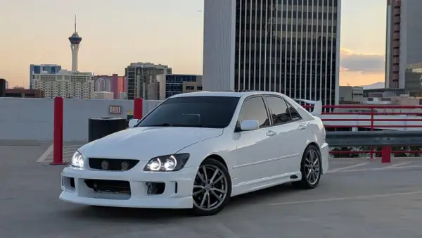 Biała Toyota Altezza z tuningowym body kitem i spojlerem na dachu parkingu, w tle wieżowce i wieża widokowa.