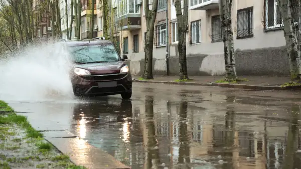 Ciemnobordowy samochód wpada w kałużę, powodując potężny rozbryzg wody. Niebezpieczny aquaplaning na mokrej ulicy.