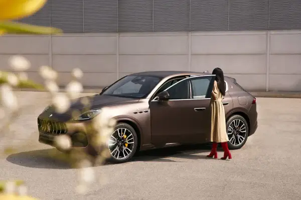 Elegancka kobieta w beżowej sukience i czerwonych kozakach wsiada do brązowego Maserati SUV.
