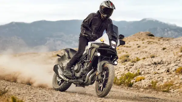 Motocyklista w kasku i kurtce pędzi na motocyklu Honda CB500X po piaszczystej drodze, wzbijając tumany kurzu.