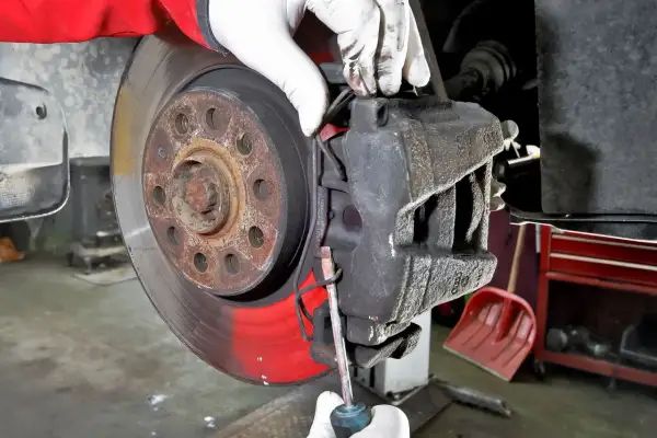 Mechanik w białych rękawiczkach dokonuje wymiany klocków hamulcowych, używając śrubokręta do podważenia starego klocka.
